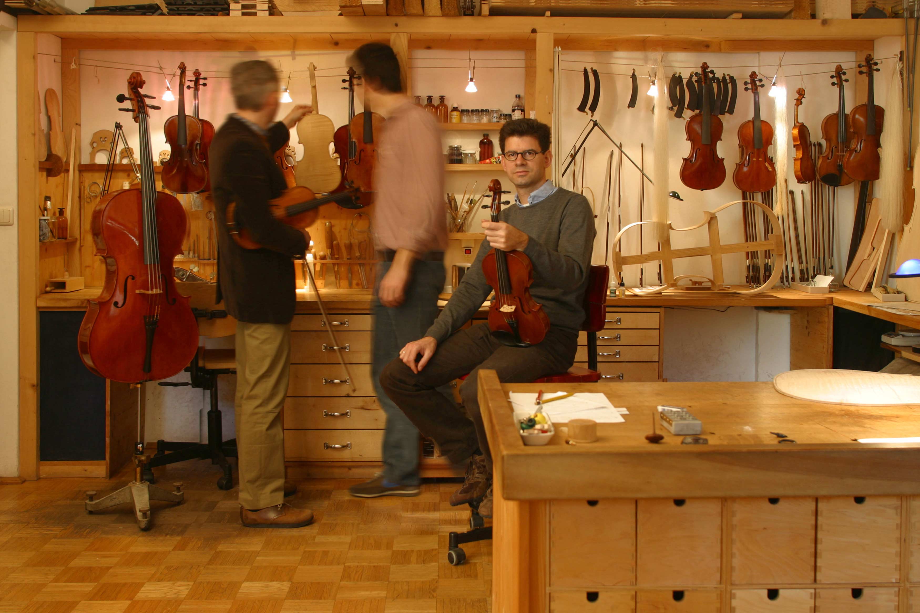 Inside violin maker's workshop. Violin maker sits beside workbench and holds violin in his hand. 2 persons standing behind him and looking at instruments.
