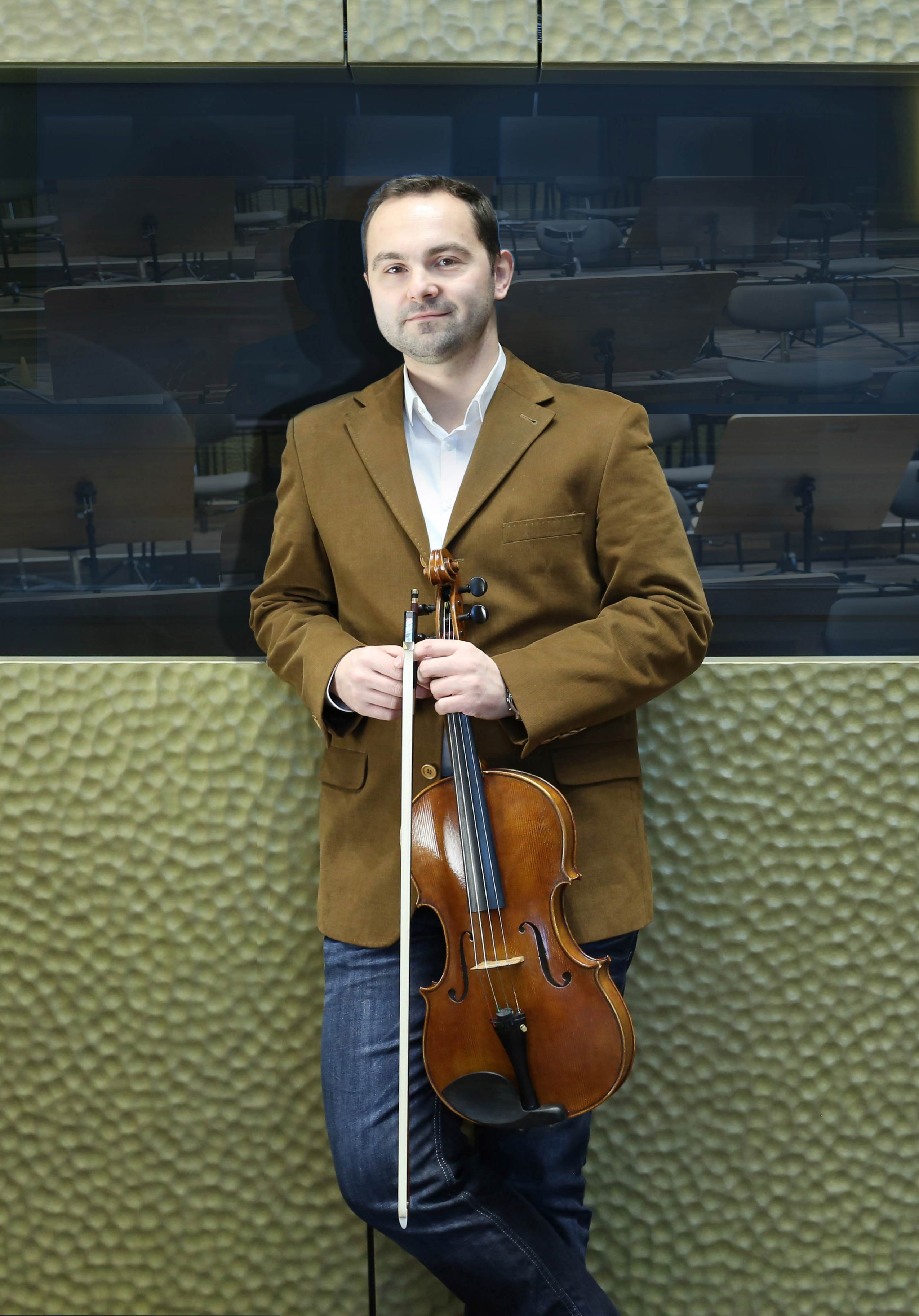 A man stands in front of a wall of a concert hall. He holds a viola in his hand.